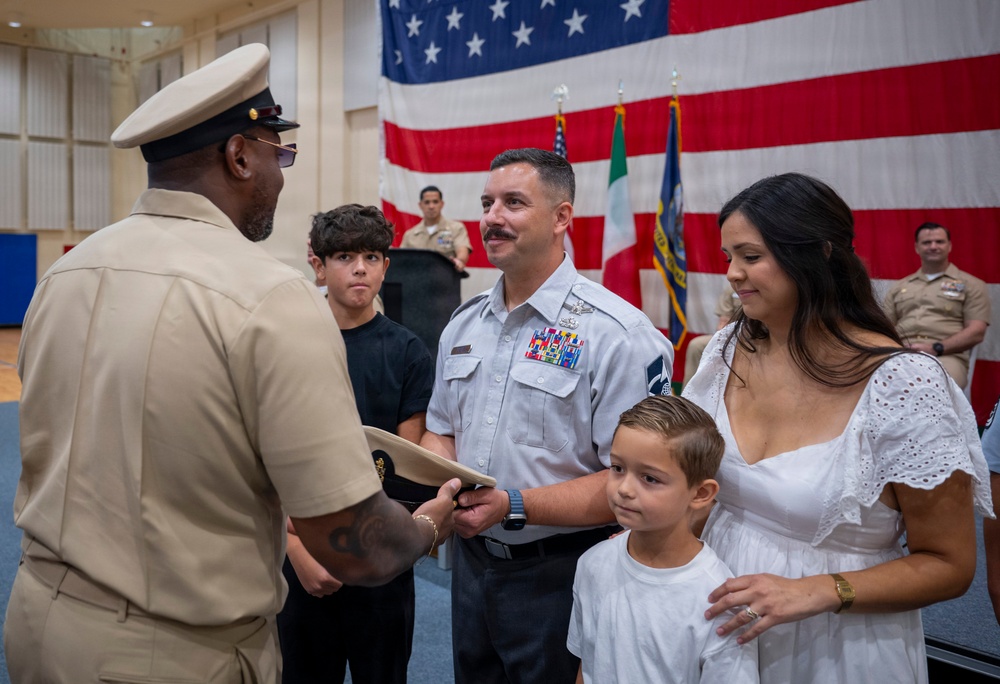 A U.S. Navy military ceremony is taking place in front of a large American flag, with additional flags visible in the background. A senior enlisted sailor in khaki uniform is presenting what appears to be a cover (hat) or certificate to a uniformed service member accompanied by his family. The occasion appears to be a promotion, retirement, or award ceremony.