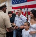 A U.S. Navy military ceremony is taking place in front of a large American flag, with additional flags visible in the background. A senior enlisted sailor in khaki uniform is presenting what appears to be a cover (hat) or certificate to a uniformed service member accompanied by his family. The occasion appears to be a promotion, retirement, or award ceremony.