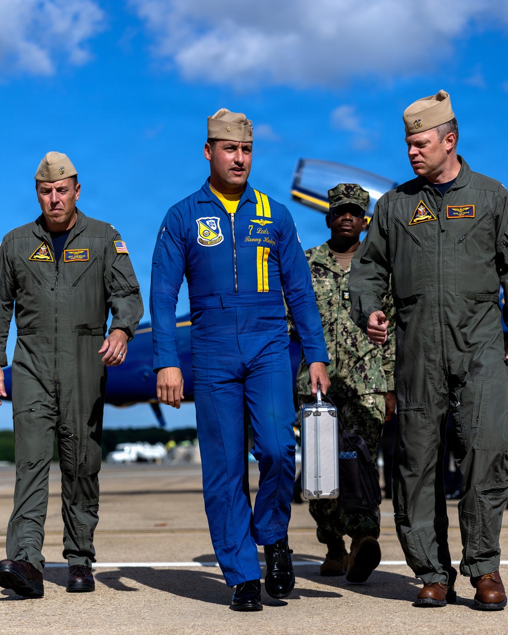 Four military personnel are walking across an airfield tarmac, with a Blue Angels aircraft visible in the background. The central figure is wearing the distinctive blue and yellow flight suit of the U.S. Navy Blue Angels demonstration team and is carrying a silver metal briefcase. The others are wearing standard olive/sage green military flight suits with garrison caps.