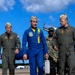 Four military personnel are walking across an airfield tarmac, with a Blue Angels aircraft visible in the background. The central figure is wearing the distinctive blue and yellow flight suit of the U.S. Navy Blue Angels demonstration team and is carrying a silver metal briefcase. The others are wearing standard olive/sage green military flight suits with garrison caps.