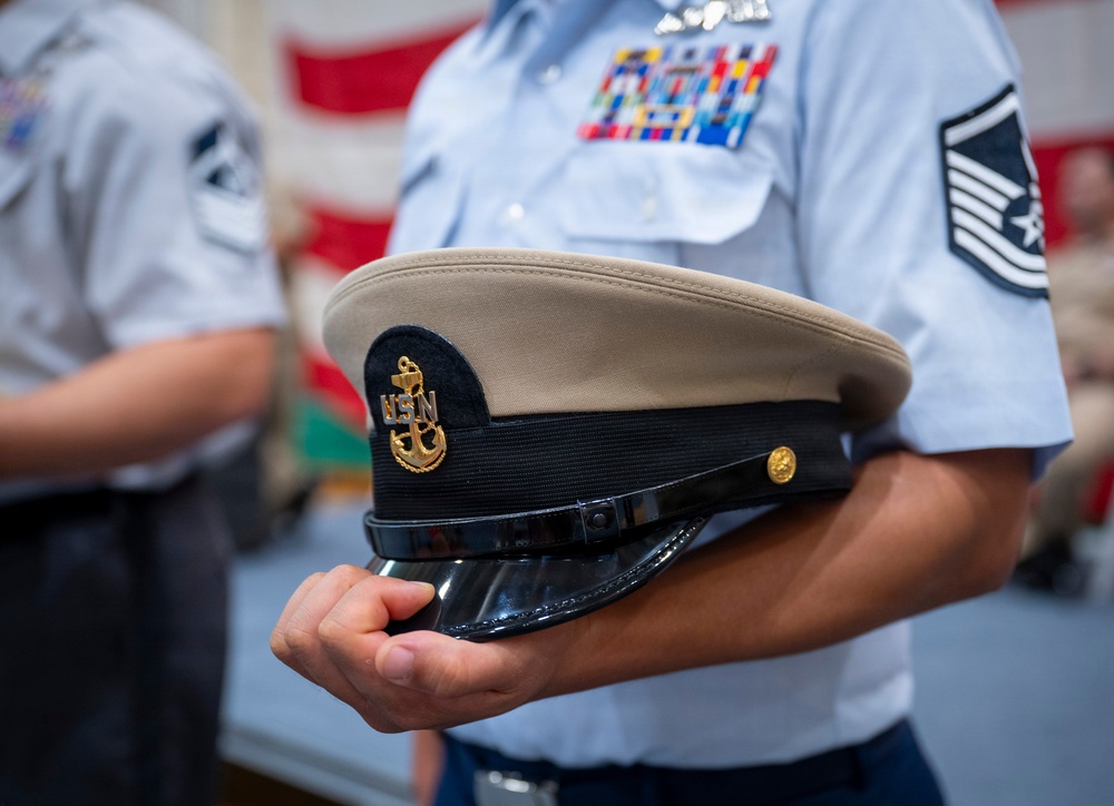 A person in a light blue U.S. Air Force uniform is holding a U.S. Navy (USN) chief petty officer's combination cover (dress cap). The Air Force member displays multiple service ribbons and a Senior Master Sergeant or Chief Master Sergeant rank insignia on the sleeve. The setting appears to be an indoor military ceremony or formal event, with an American flag visible in the background.