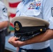 A person in a light blue U.S. Air Force uniform is holding a U.S. Navy (USN) chief petty officer's combination cover (dress cap). The Air Force member displays multiple service ribbons and a Senior Master Sergeant or Chief Master Sergeant rank insignia on the sleeve. The setting appears to be an indoor military ceremony or formal event, with an American flag visible in the background.