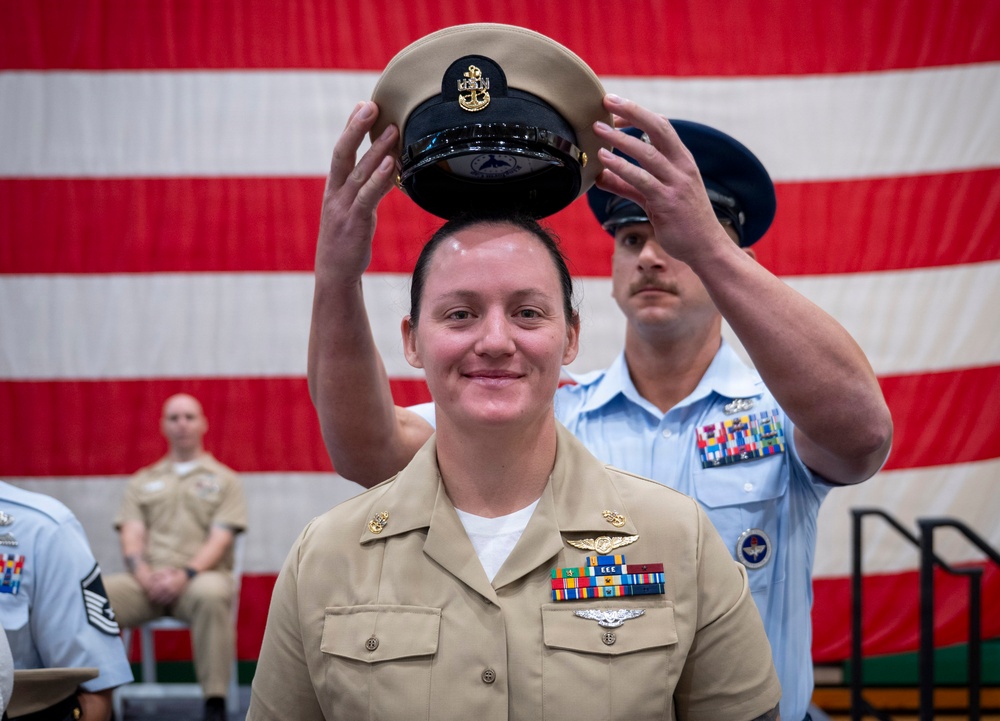A U.S. Navy Chief Petty Officer promotion ceremony is taking place in front of a large American flag backdrop. A female sailor is having the distinctive khaki Chief Petty Officer cover (hat) placed on her head, marking her advancement in rank. A male service member in a light blue uniform, likely Air Force, is performing the hat-placing honors.