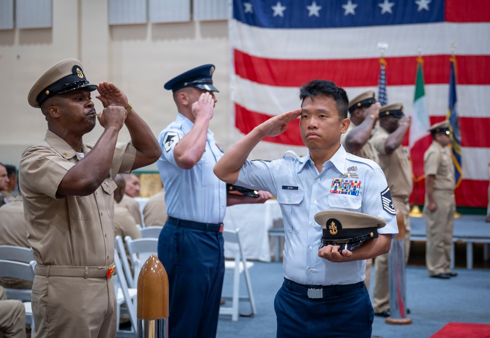 A formal military ceremony is taking place indoors in front of a large American flag. Multiple service members from different branches — including the U.S. Navy and U.S. Air Force — are rendering salutes simultaneously. The foreground figure in an Air Force light blue uniform holds a Navy cover (hat) while saluting, suggesting a joint-service recognition or retirement/promotion event.