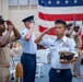 A formal military ceremony is taking place indoors in front of a large American flag. Multiple service members from different branches — including the U.S. Navy and U.S. Air Force — are rendering salutes simultaneously. The foreground figure in an Air Force light blue uniform holds a Navy cover (hat) while saluting, suggesting a joint-service recognition or retirement/promotion event.
