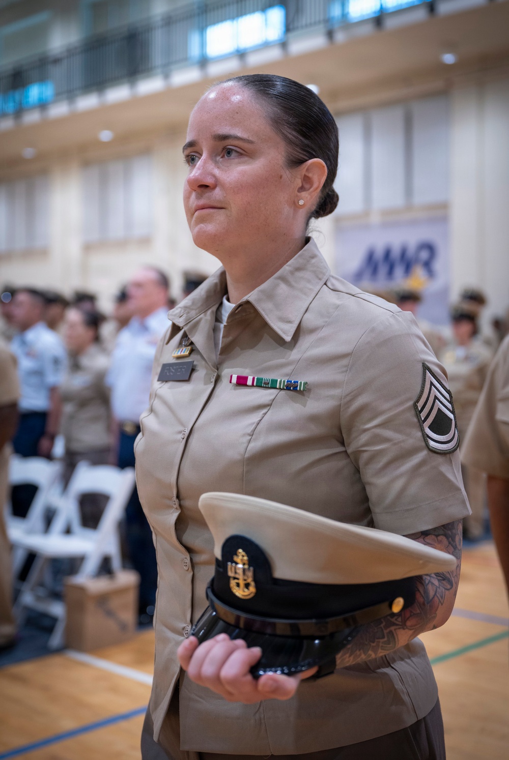 A female U.S. Navy enlisted service member in tan/khaki uniform stands at attention during what appears to be a formal military ceremony held indoors in a gymnasium or large hall. She is holding a combination cover (dress cap) with a gold anchor device and is wearing service ribbons and a rank insignia consistent with a Senior Chief Petty Officer on her right sleeve. The setting includes an MWR (Morale, Welfare, and Recreation) banner in the background with other uniformed personnel present.