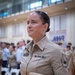 A female U.S. Navy enlisted service member in tan/khaki uniform stands at attention during what appears to be a formal military ceremony held indoors in a gymnasium or large hall. She is holding a combination cover (dress cap) with a gold anchor device and is wearing service ribbons and a rank insignia consistent with a Senior Chief Petty Officer on her right sleeve. The setting includes an MWR (Morale, Welfare, and Recreation) banner in the background with other uniformed personnel present.