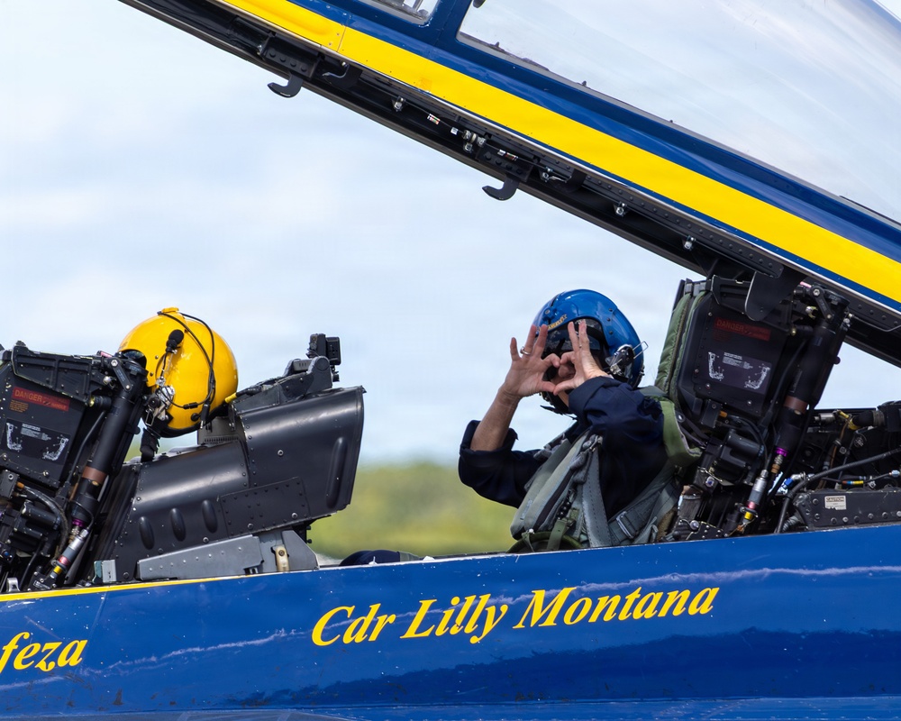 A two-seat military jet aircraft, painted in the distinctive blue and gold colors of the U.S. Navy Blue Angels, is shown on the ground with its canopy open. The rear cockpit occupant, identified by fuselage text as 'Cdr Lilly Montana,' is wearing a blue flight helmet and making a heart shape with their hands toward the camera. The front cockpit contains a yellow helmet resting on the ejection seat.