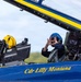 A two-seat military jet aircraft, painted in the distinctive blue and gold colors of the U.S. Navy Blue Angels, is shown on the ground with its canopy open. The rear cockpit occupant, identified by fuselage text as 'Cdr Lilly Montana,' is wearing a blue flight helmet and making a heart shape with their hands toward the camera. The front cockpit contains a yellow helmet resting on the ejection seat.