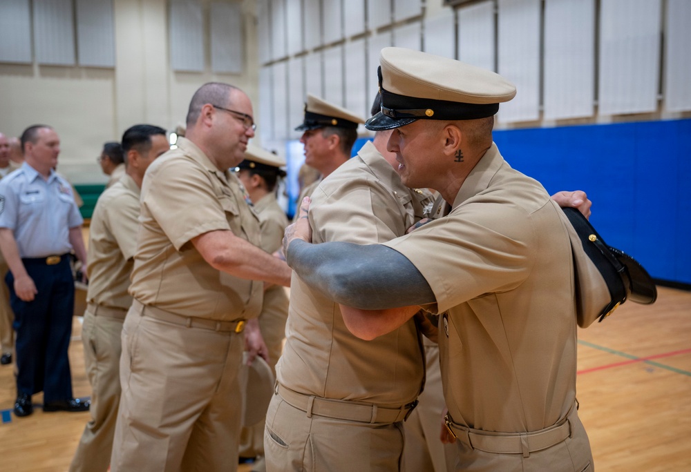 A group of U.S. Navy personnel in tan/khaki service uniforms are gathered in what appears to be a gymnasium or indoor facility. Two sailors in the foreground are embracing, suggesting a congratulatory or farewell moment, likely during a ceremony or promotion event. Additional Navy personnel and at least one individual in a lighter blue uniform are visible in the background.