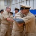 A group of U.S. Navy personnel in tan/khaki service uniforms are gathered in what appears to be a gymnasium or indoor facility. Two sailors in the foreground are embracing, suggesting a congratulatory or farewell moment, likely during a ceremony or promotion event. Additional Navy personnel and at least one individual in a lighter blue uniform are visible in the background.
