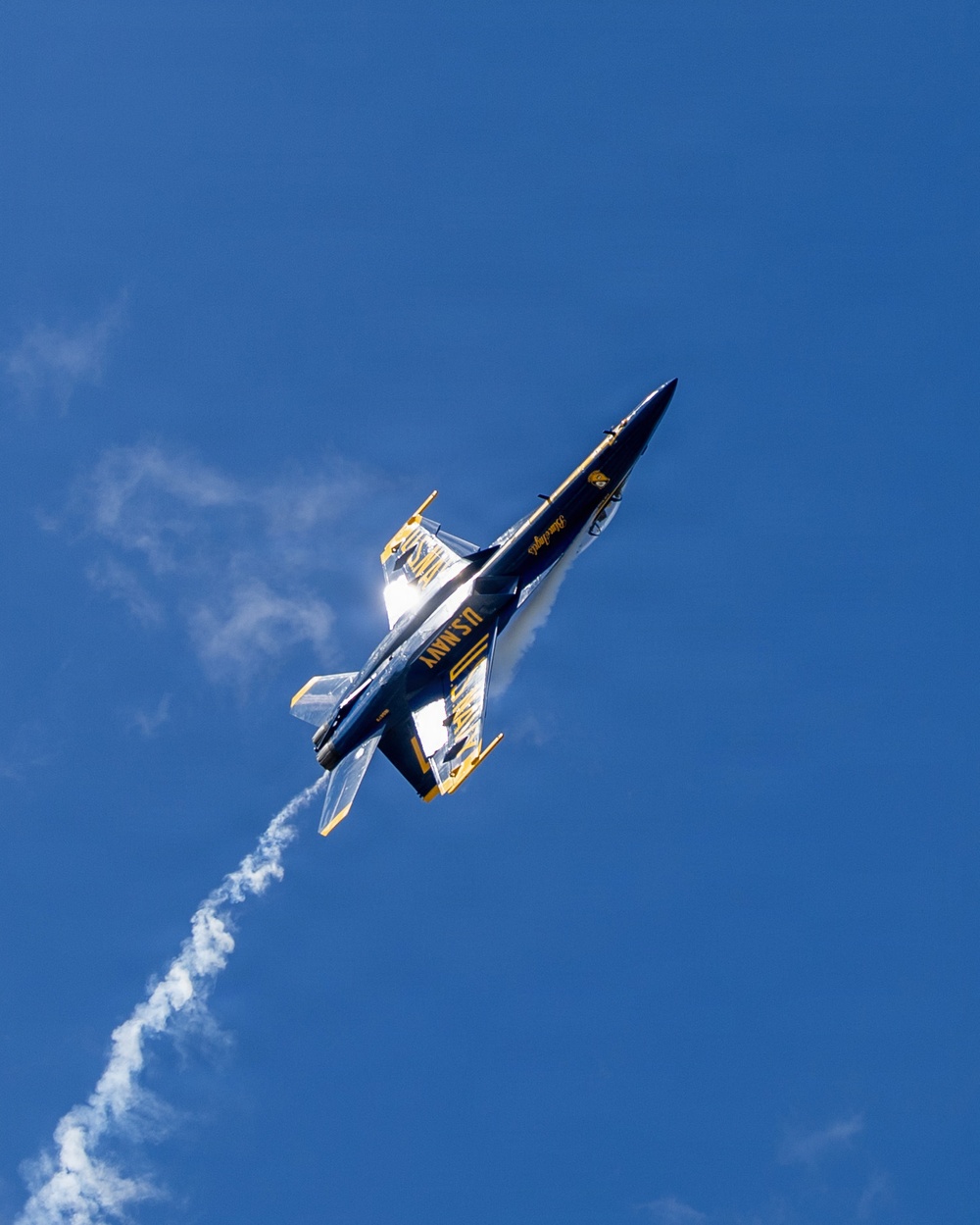 A U.S. Navy Blue Angels F/A-18 Hornet jet is captured in a steep climbing maneuver against a clear blue sky. The aircraft is photographed from below, showing its distinctive navy blue and gold livery with 'U.S. NAVY' lettering clearly visible on the underside. A white smoke trail extends behind the aircraft, indicating an aerobatic performance.
