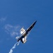 A U.S. Navy Blue Angels F/A-18 Hornet jet is captured in a steep climbing maneuver against a clear blue sky. The aircraft is photographed from below, showing its distinctive navy blue and gold livery with 'U.S. NAVY' lettering clearly visible on the underside. A white smoke trail extends behind the aircraft, indicating an aerobatic performance.