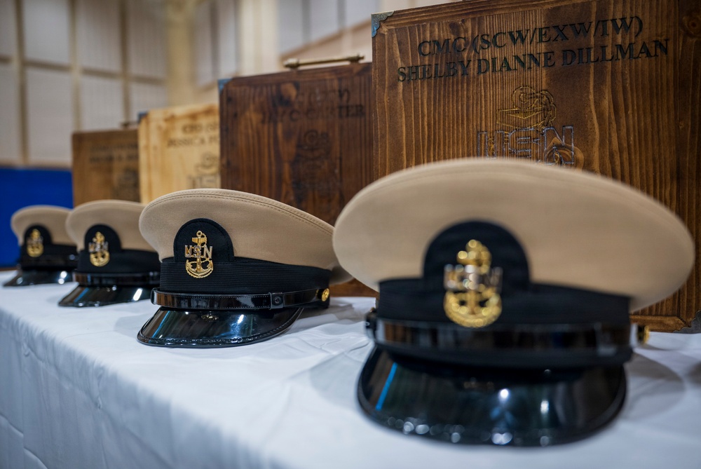 The image shows a ceremonial display of U.S. Navy Chief Petty Officer combination covers (dress caps) arranged on a white-cloth-covered table. Behind each cap is a personalized wooden shadow box or presentation case, with the foremost one inscribed 'CMC(SCW/EXW/TW) SHELBY DIANNE DILLMAN'. This appears to be a Navy Chief Petty Officer pinning or advancement ceremony.