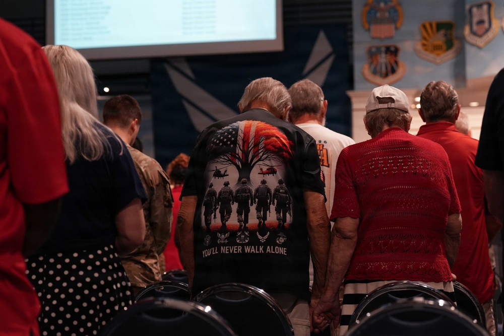 A group of people stand with bowed heads in what appears to be a military memorial or veterans' ceremony. The central figure wears a patriotic t-shirt reading 'You'll Never Walk Alone' featuring soldiers, helicopters, and an American flag design. Military insignia and branch emblems are visible on the wall in the background.