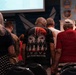 A group of people stand with bowed heads in what appears to be a military memorial or veterans' ceremony. The central figure wears a patriotic t-shirt reading 'You'll Never Walk Alone' featuring soldiers, helicopters, and an American flag design. Military insignia and branch emblems are visible on the wall in the background.