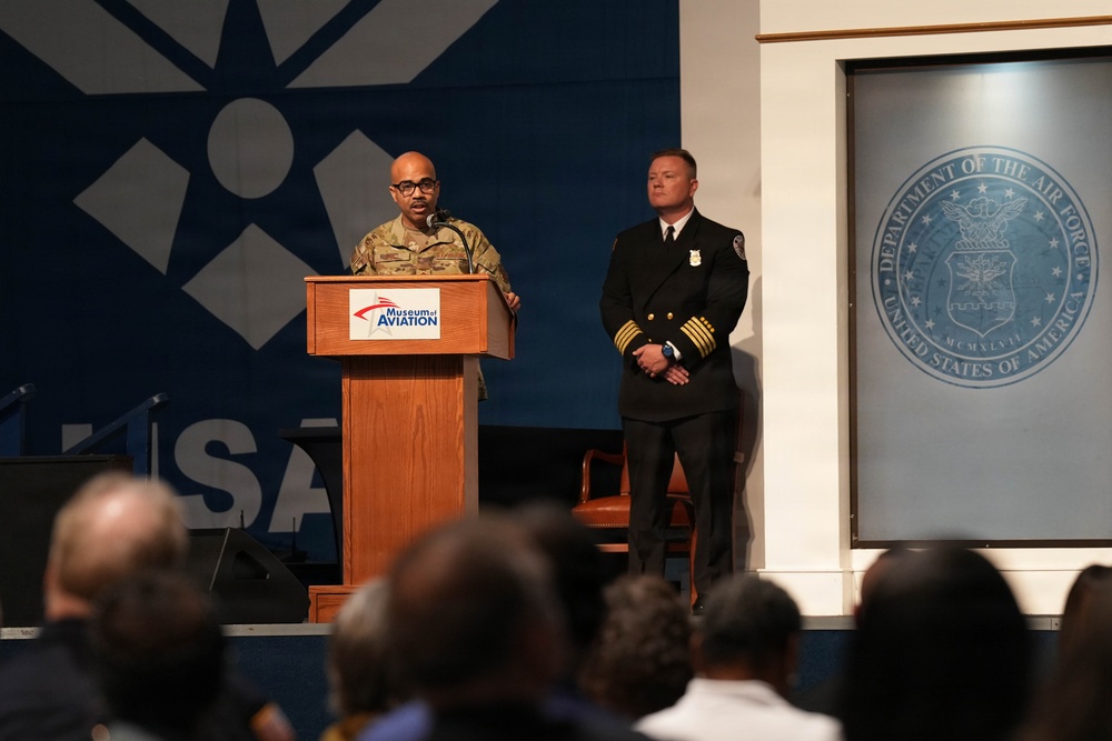 A formal ceremony or event is taking place at the Museum of Aviation, with a person in military camouflage uniform speaking at a podium. A second individual in a dark formal uniform with gold insignia stands to the right. The Department of the Air Force seal is prominently displayed on the wall, and an audience is visible in the foreground.
