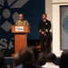 A formal ceremony or event is taking place at the Museum of Aviation, with a person in military camouflage uniform speaking at a podium. A second individual in a dark formal uniform with gold insignia stands to the right. The Department of the Air Force seal is prominently displayed on the wall, and an audience is visible in the foreground.