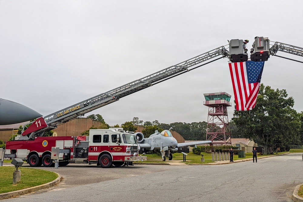 A U.S. Air Force fire truck (Unit 11) has its aerial ladder fully extended, suspending a large American flag over a roadway in what appears to be a ceremonial display. The scene is set on a military installation, likely an Air Force base, with a static display aircraft (A-10 Thunderbolt II) and a control tower visible in the background. Two personnel are standing on the ground near the right side of the frame, suggesting a formal or commemorative event.