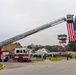 A U.S. Air Force fire truck (Unit 11) has its aerial ladder fully extended, suspending a large American flag over a roadway in what appears to be a ceremonial display. The scene is set on a military installation, likely an Air Force base, with a static display aircraft (A-10 Thunderbolt II) and a control tower visible in the background. Two personnel are standing on the ground near the right side of the frame, suggesting a formal or commemorative event.