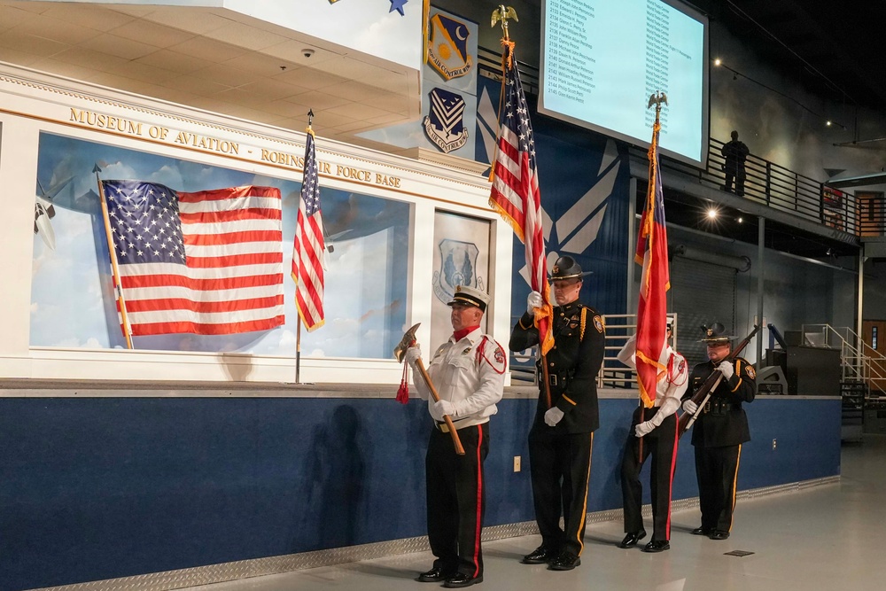 A color guard ceremony is taking place inside the Museum of Aviation at Robins Air Force Base. Four uniformed personnel — appearing to represent fire and law enforcement services — are presenting flags including the U.S. flag and what appears to be a state flag. A projection screen displaying names is visible in the background, suggesting a memorial or recognition event.