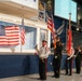 A color guard ceremony is taking place inside the Museum of Aviation at Robins Air Force Base. Four uniformed personnel — appearing to represent fire and law enforcement services — are presenting flags including the U.S. flag and what appears to be a state flag. A projection screen displaying names is visible in the background, suggesting a memorial or recognition event.