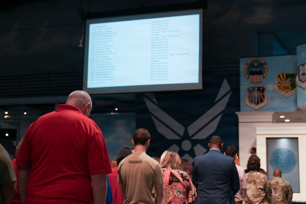 A group of people are gathered in what appears to be a United States Air Force facility or museum, facing a large projection screen displaying a list of names or text. The room features prominent U.S. Air Force insignia and military unit emblems on the walls. The gathering appears to be a formal or ceremonial event based on the mix of civilian and military attire.