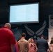 A group of people are gathered in what appears to be a United States Air Force facility or museum, facing a large projection screen displaying a list of names or text. The room features prominent U.S. Air Force insignia and military unit emblems on the walls. The gathering appears to be a formal or ceremonial event based on the mix of civilian and military attire.