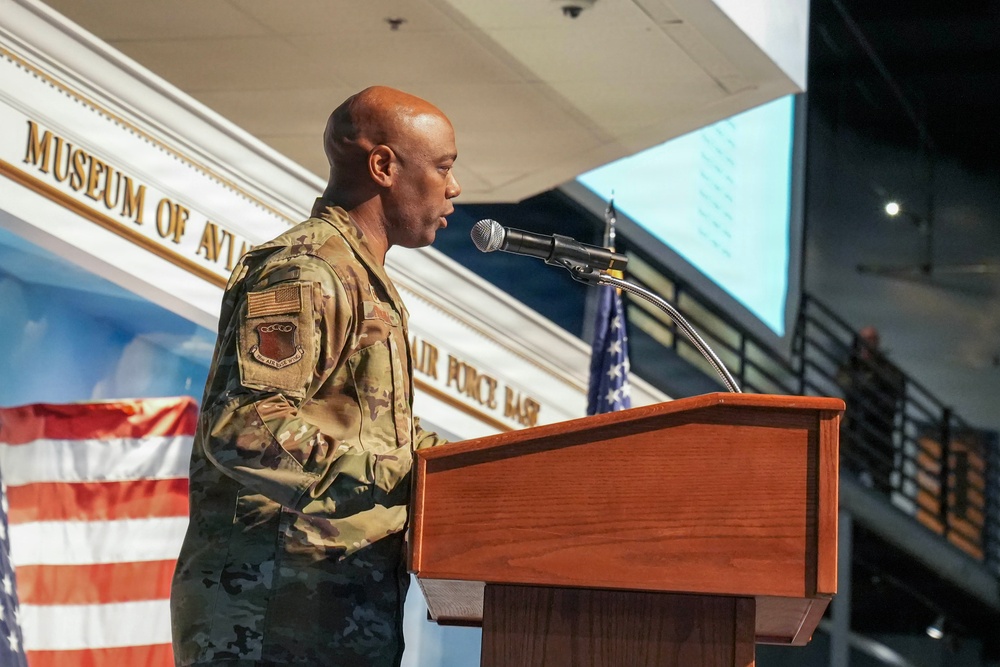 A U.S. Air Force military officer in OCP (Operational Camouflage Pattern) uniform is speaking at a podium with a microphone at the Museum of Aviation. The event appears to be an official military ceremony or address, taking place inside the museum facility. An American flag and what appears to be a unit flag are visible in the background.