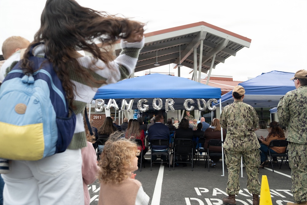 An outdoor ceremony is taking place in a parking lot at what appears to be the Bangor CDC (Child Development Center), indicated by silver balloon letters under a blue canopy tent. Military personnel in Navy Working Uniform (NWU) camouflage are present alongside civilians, including families with children. The event appears to be a dedication, ribbon-cutting, or opening ceremony for the facility.