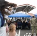 An outdoor ceremony is taking place in a parking lot at what appears to be the Bangor CDC (Child Development Center), indicated by silver balloon letters under a blue canopy tent. Military personnel in Navy Working Uniform (NWU) camouflage are present alongside civilians, including families with children. The event appears to be a dedication, ribbon-cutting, or opening ceremony for the facility.