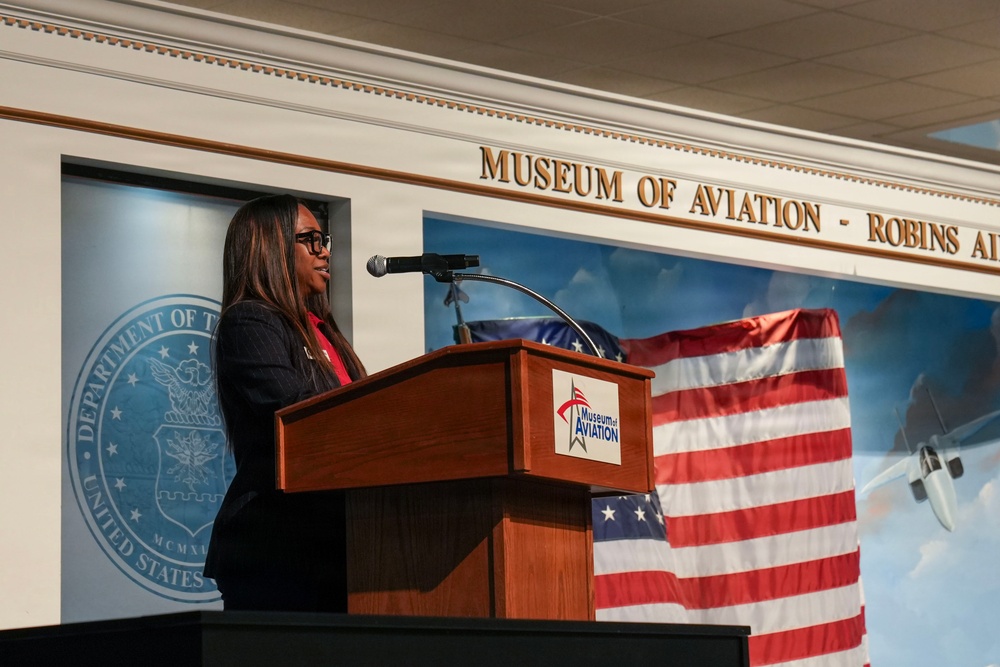 A woman is delivering a speech at a podium inside the Museum of Aviation at Robins Air Force Base. The setting appears to be a formal event, with an American flag and a Department of the Air Force seal visible in the background. The podium bears the Museum of Aviation logo.