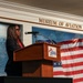 A woman is delivering a speech at a podium inside the Museum of Aviation at Robins Air Force Base. The setting appears to be a formal event, with an American flag and a Department of the Air Force seal visible in the background. The podium bears the Museum of Aviation logo.
