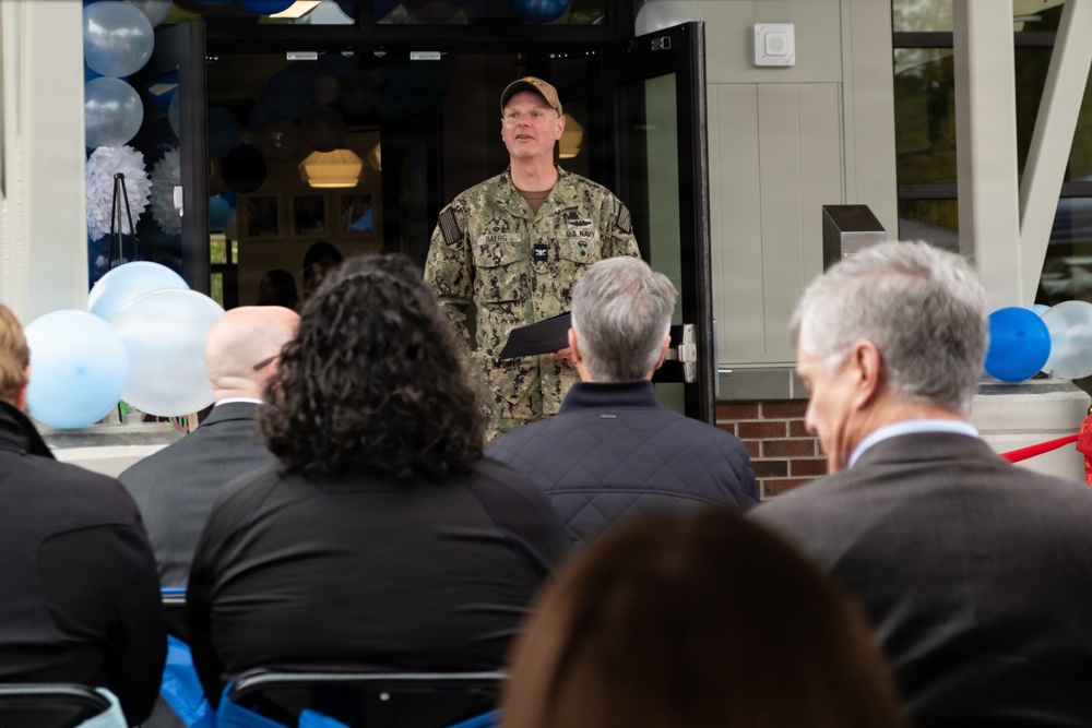 A U.S. Navy officer in camouflage uniform is delivering a speech at what appears to be a ribbon-cutting or dedication ceremony outside a building. The speaker stands in front of an open glass door decorated with blue and white balloons, addressing a seated audience of civilians in formal attire. The name tape on the uniform appears to read 'DAERG' and a U.S. Navy insignia is visible.