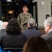 A U.S. Navy officer in camouflage uniform is delivering a speech at what appears to be a ribbon-cutting or dedication ceremony outside a building. The speaker stands in front of an open glass door decorated with blue and white balloons, addressing a seated audience of civilians in formal attire. The name tape on the uniform appears to read 'DAERG' and a U.S. Navy insignia is visible.