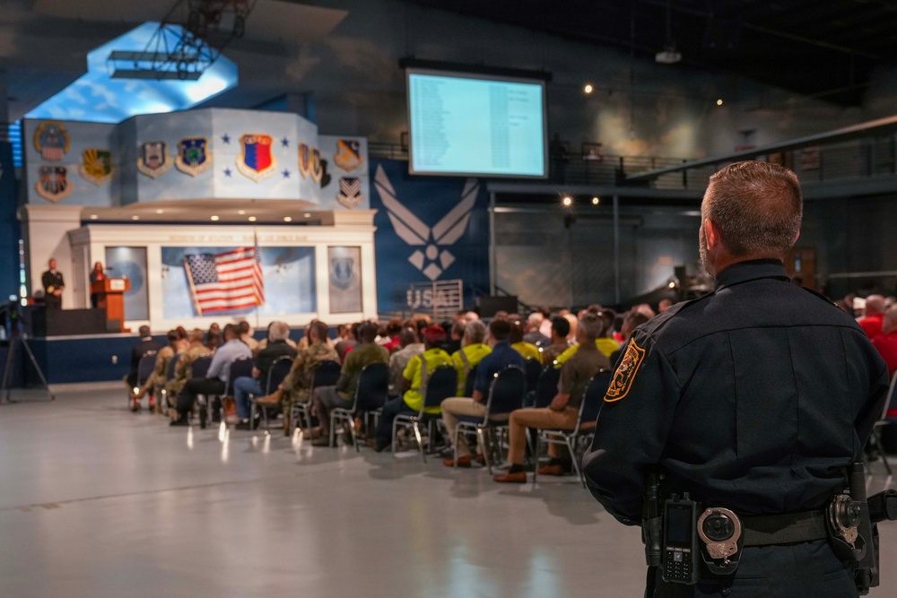 A formal ceremony or event is taking place inside what appears to be a United States Air Force museum or hangar, identifiable by the USAF logo and military insignia displayed on the stage backdrop. A law enforcement officer in a dark uniform with a duty belt stands in the foreground observing the seated audience. A speaker addresses the crowd from a podium on a decorated stage featuring an American flag and multiple Air Force command patches.