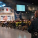 A formal ceremony or event is taking place inside what appears to be a United States Air Force museum or hangar, identifiable by the USAF logo and military insignia displayed on the stage backdrop. A law enforcement officer in a dark uniform with a duty belt stands in the foreground observing the seated audience. A speaker addresses the crowd from a podium on a decorated stage featuring an American flag and multiple Air Force command patches.