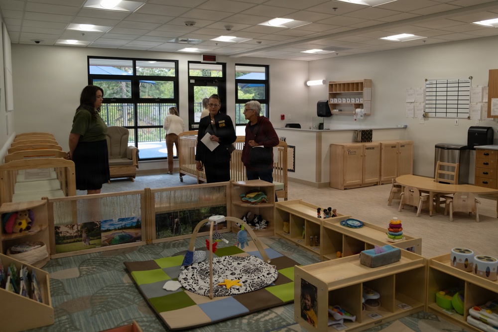 Three adults are standing and conversing in what appears to be a newly opened or recently set up infant/toddler childcare classroom. The room is well-equipped with wooden furniture, cribs, play mats, toy shelves, and learning materials typical of an early childhood education facility. The setting appears clean and modern, suggesting this may be a tour or inspection of the facility.