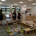 Three adults are standing and conversing in what appears to be a newly opened or recently set up infant/toddler childcare classroom. The room is well-equipped with wooden furniture, cribs, play mats, toy shelves, and learning materials typical of an early childhood education facility. The setting appears clean and modern, suggesting this may be a tour or inspection of the facility.