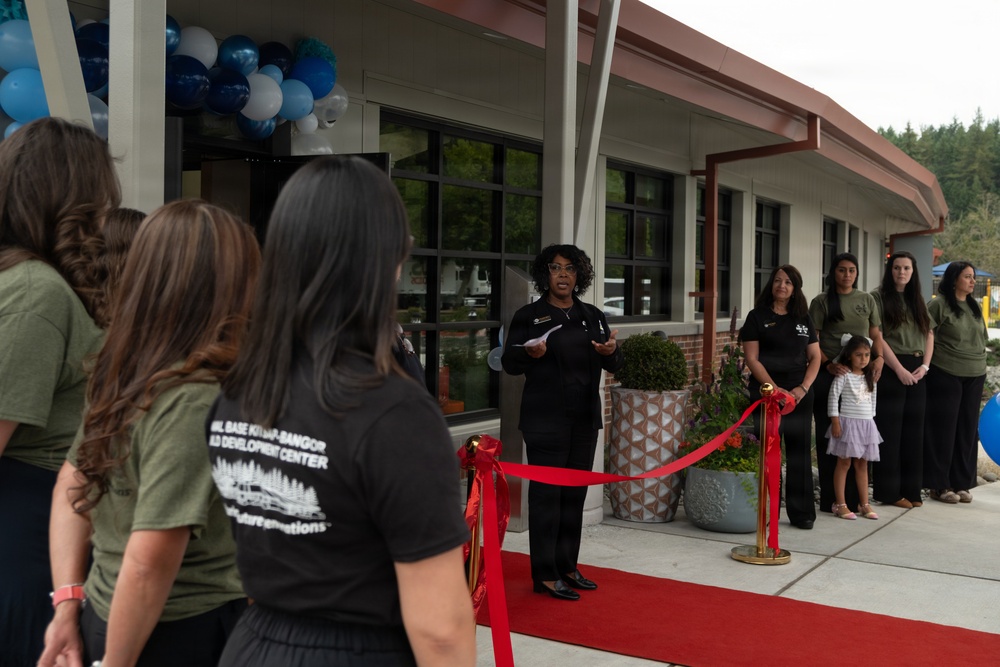 A ribbon-cutting ceremony is taking place at what appears to be the Naval Base Kitsap-Bangor Child Development Center. A woman in black attire is speaking at a podium area in front of the building, which is decorated with blue, white, and silver balloons. Staff members wearing olive green and black t-shirts are gathered on both sides of a red carpet and red ribbon barrier.