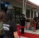 A ribbon-cutting ceremony is taking place at what appears to be the Naval Base Kitsap-Bangor Child Development Center. A woman in black attire is speaking at a podium area in front of the building, which is decorated with blue, white, and silver balloons. Staff members wearing olive green and black t-shirts are gathered on both sides of a red carpet and red ribbon barrier.