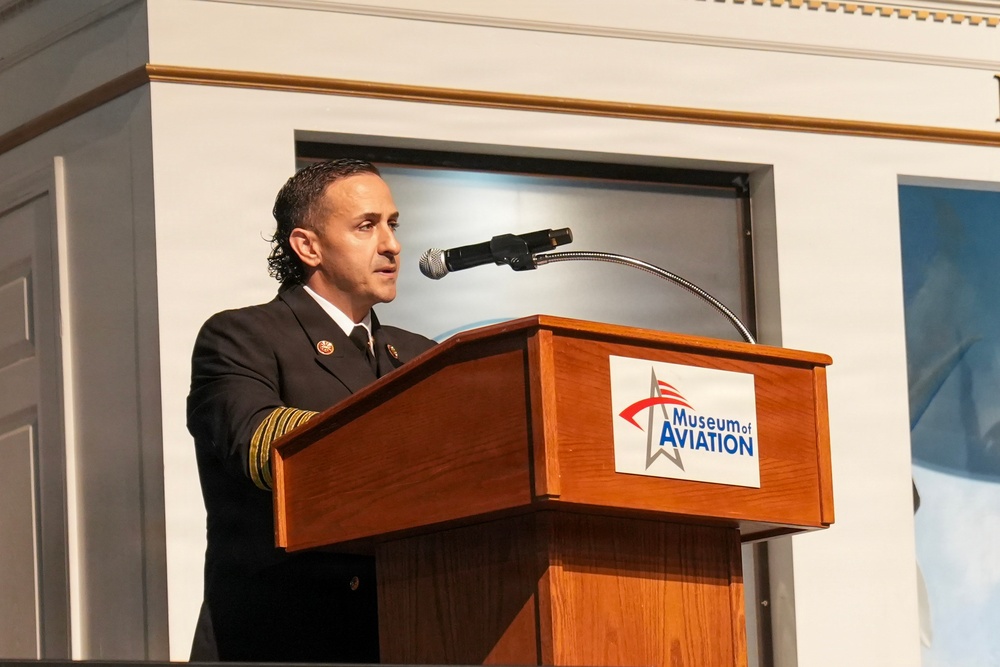 A uniformed official is speaking at a podium bearing the 'Museum of Aviation' logo. The speaker appears to be wearing a dark dress uniform with gold sleeve stripes and insignia, suggesting a military or fire service rank. The setting appears to be a formal event or ceremony held at or associated with the Museum of Aviation.