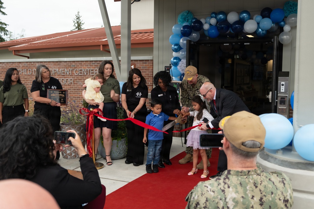 A ribbon-cutting ceremony is taking place at a Child Development Center, likely on a military installation based on the Navy uniforms visible. Children are participating in cutting a red ribbon with large scissors, accompanied by adults in both civilian and military attire. The entrance is decorated with a balloon arch in blue, teal, and silver colors.