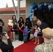 A ribbon-cutting ceremony is taking place at a Child Development Center, likely on a military installation based on the Navy uniforms visible. Children are participating in cutting a red ribbon with large scissors, accompanied by adults in both civilian and military attire. The entrance is decorated with a balloon arch in blue, teal, and silver colors.