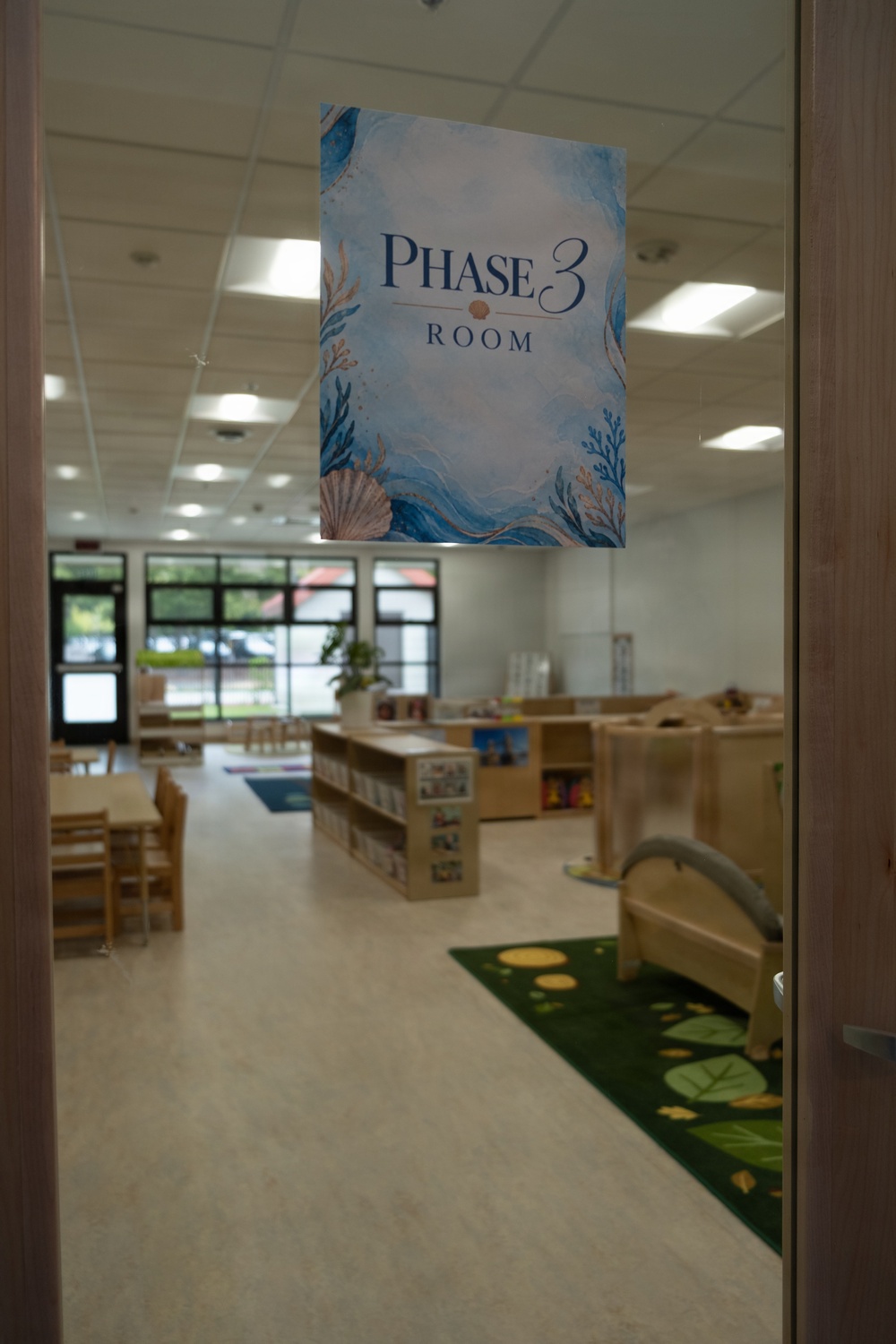 The image shows a view through a glass door into a children's classroom or daycare room labeled 'Phase 3 Room.' The sign features an ocean/underwater theme with seashells and coral decorations. The room appears to be empty of people and is well-furnished with child-sized furniture, bookshelves, and colorful rugs.