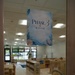 The image shows a view through a glass door into a children's classroom or daycare room labeled 'Phase 3 Room.' The sign features an ocean/underwater theme with seashells and coral decorations. The room appears to be empty of people and is well-furnished with child-sized furniture, bookshelves, and colorful rugs.