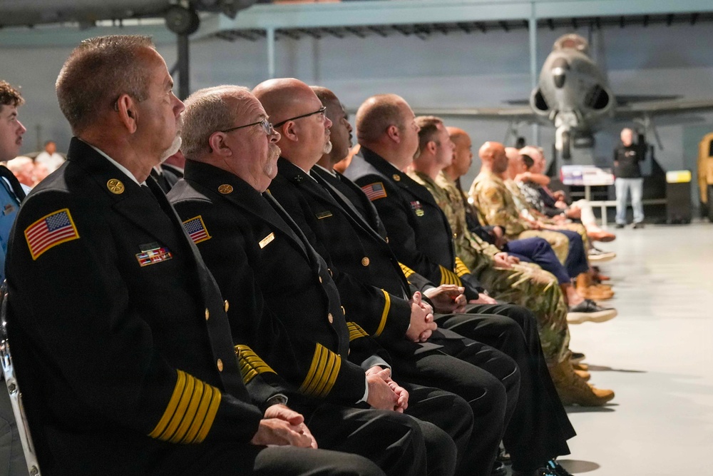 A formal ceremony is taking place inside what appears to be an aviation museum or aircraft hangar, with a row of uniformed officials seated as an audience. The attendees include individuals in dark navy/fire department dress uniforms with gold insignia and American flag patches, as well as military personnel in camouflage uniforms. A speaker or presenter is visible in the background near a displayed aircraft.