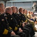 A formal ceremony is taking place inside what appears to be an aviation museum or aircraft hangar, with a row of uniformed officials seated as an audience. The attendees include individuals in dark navy/fire department dress uniforms with gold insignia and American flag patches, as well as military personnel in camouflage uniforms. A speaker or presenter is visible in the background near a displayed aircraft.