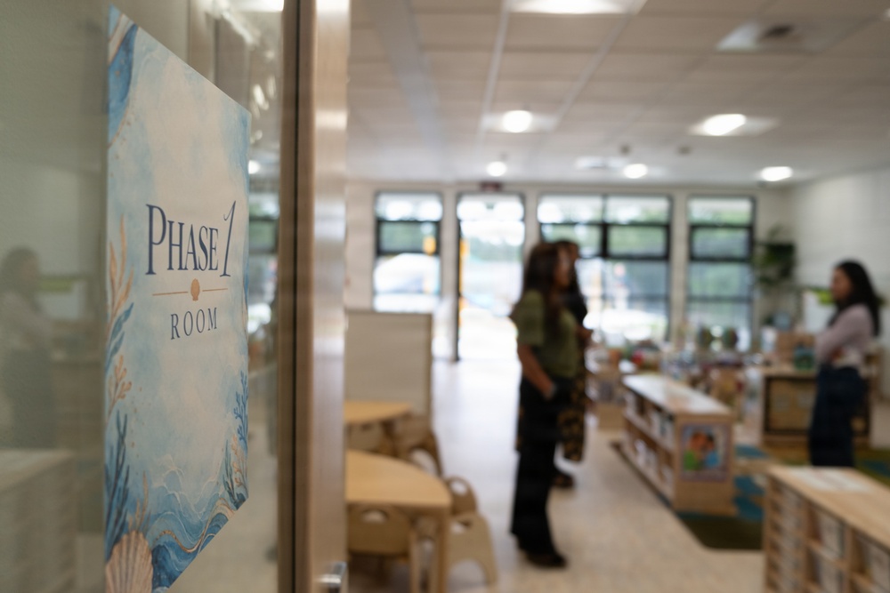 The image shows the entrance to a 'Phase 1 Room,' likely a classroom or early childhood education space. A decorative ocean-themed sign is affixed to a glass door in the foreground, while two adults stand inside the well-lit room in the background. The room contains child-sized furniture, bookshelves, and educational materials.