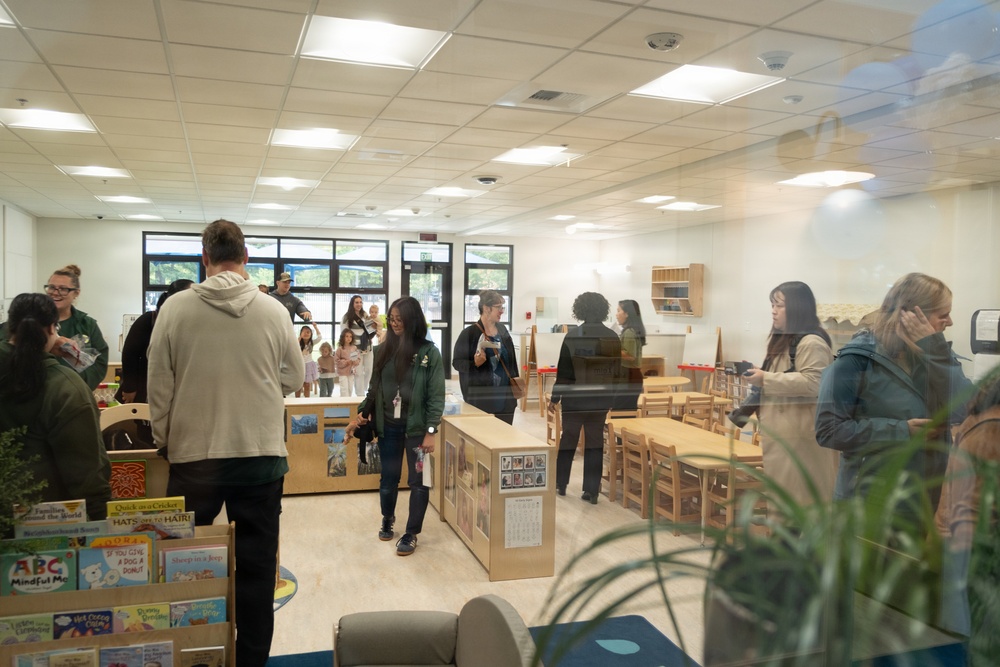 The image shows a group of adults gathered inside what appears to be a children's classroom or early education facility, likely during an open house or tour event. The room is well-lit with fluorescent ceiling lights and features child-sized furniture, educational displays, and a bookshelf with children's books. The photo is taken through a glass window or door, causing partial reflections that slightly obscure the view.